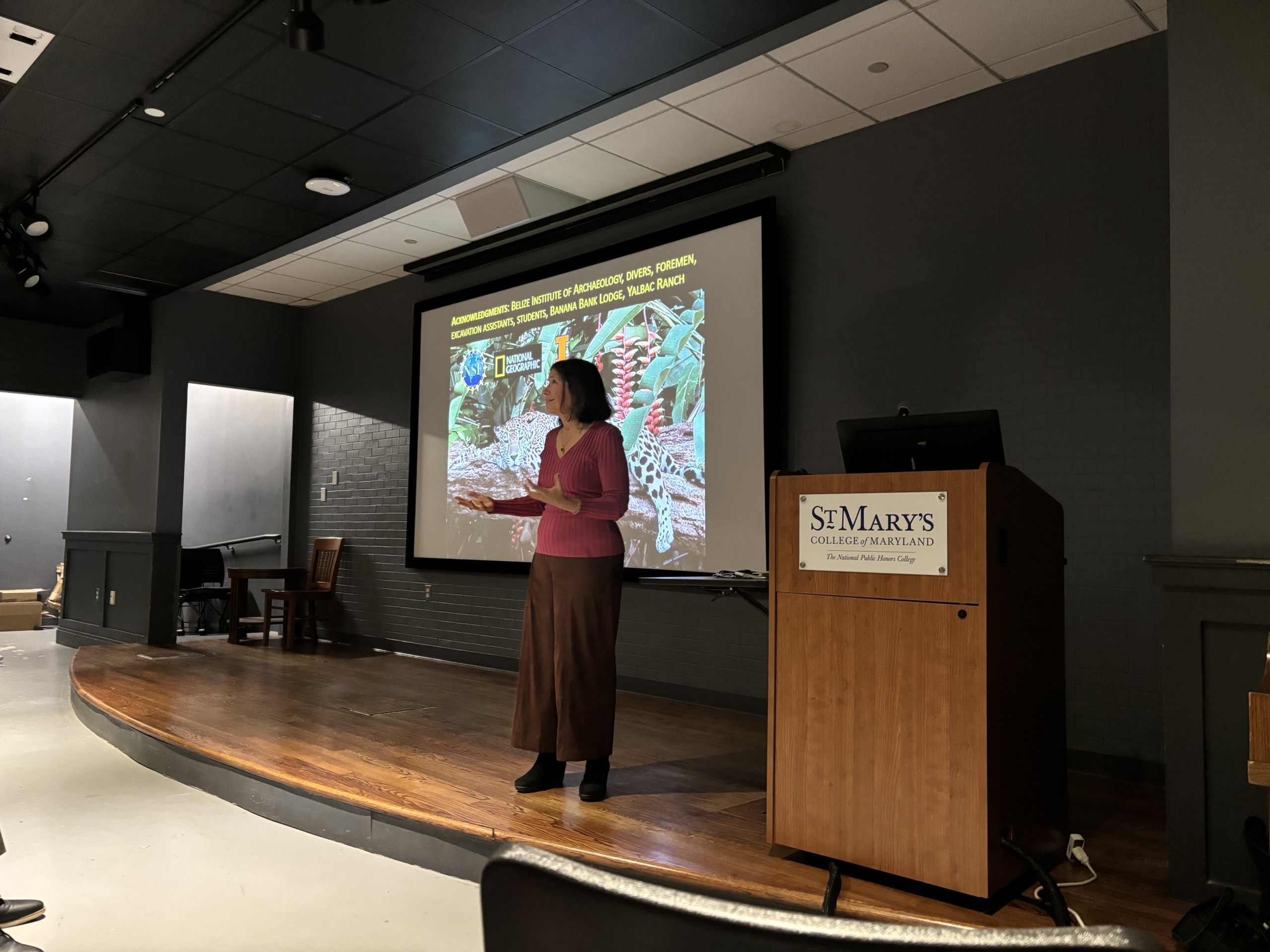 A woman stands on stage giving a presentation at St. Mary's College of Maryland, with a colorful academic slide projected behind her.