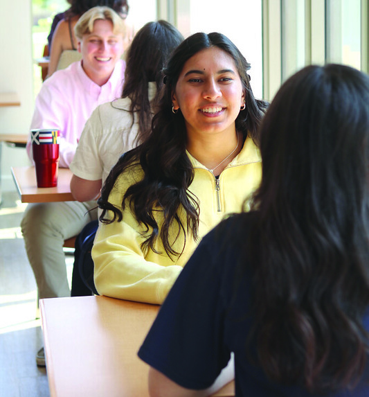 53959281193_72f5e998cf_c Several students sit in pairs at tables by a large window, engaged in conversation inside a well-lit room.