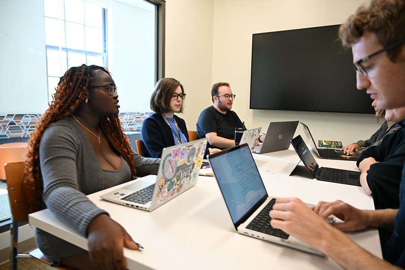 A group of people sit around a table with laptops open, engaged in discussion in a bright conference room.
