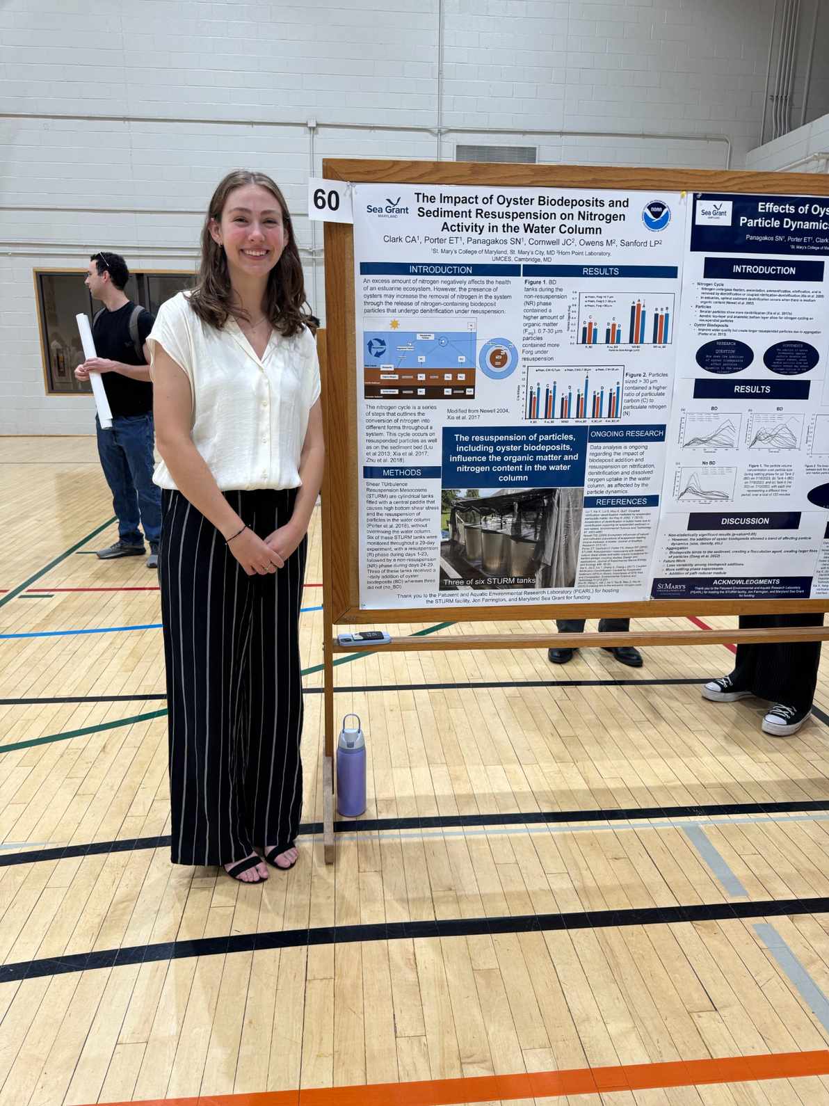 A young woman stands indoors next to a scientific poster titled "The Impact of Oyster Biodeposits and Sediment Resuspension on Nitrogen Activity in the Water Column.