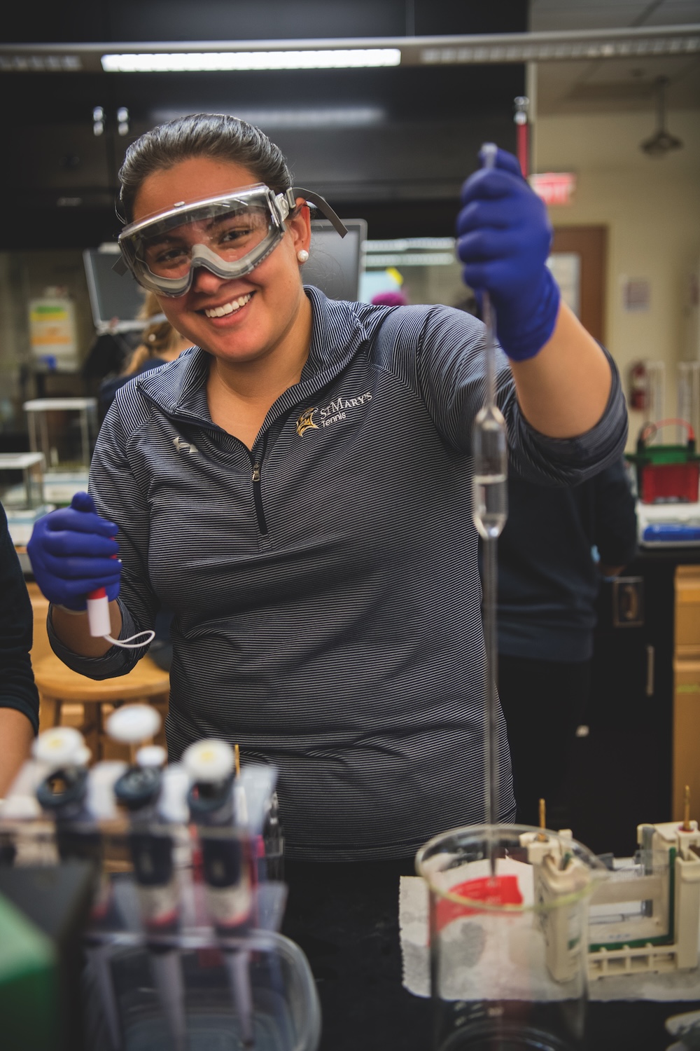 A person wearing safety goggles and gloves conducts a laboratory experiment, using a pipette and burette, and smiles at the camera.