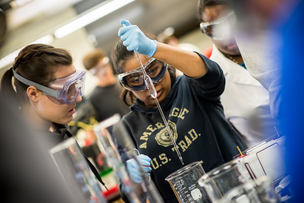 Students wearing safety goggles and gloves conduct a chemistry experiment, transferring liquid with a pipette into a beaker in a laboratory setting.