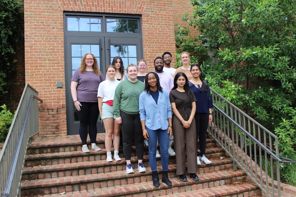 IMG_6904-scaled A group of ten people pose together on outdoor brick steps in front of a building with glass doors and greenery on the side.