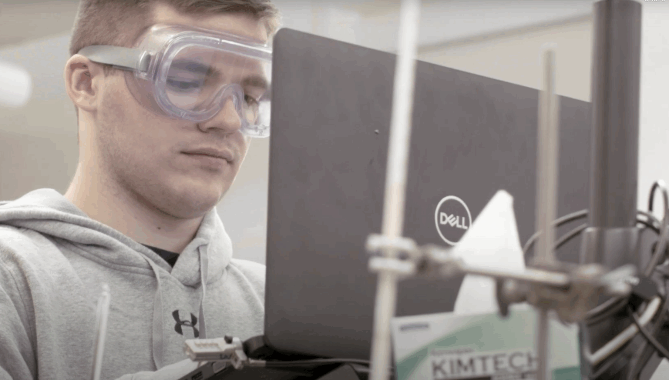 Screenshot 2025-08-06 at 9.46.22 AM A person wearing safety goggles works on a Dell laptop in a laboratory setting next to lab equipment and a box of Kimtech wipes.
