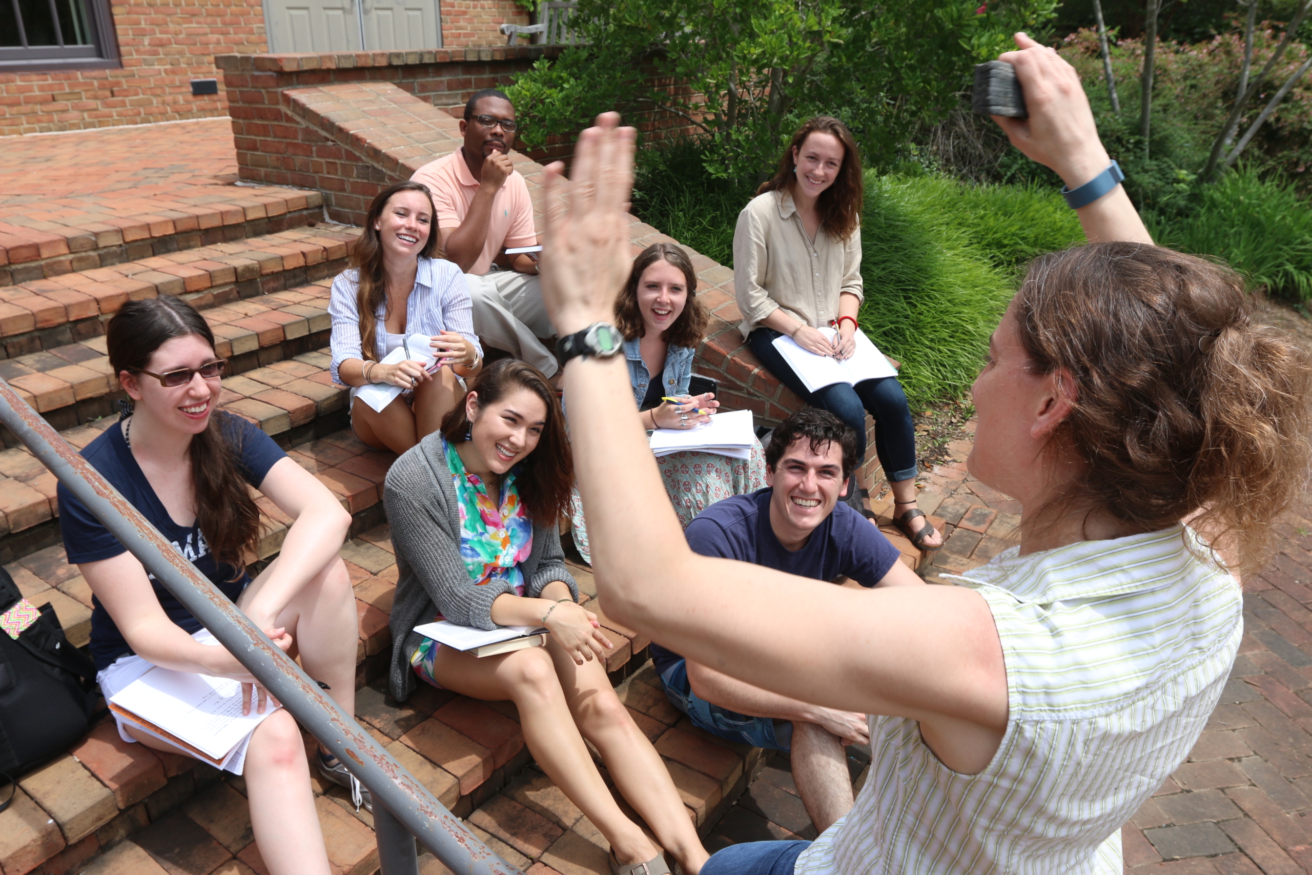 35117818476_1284a32ef3_o A group of students sit on outdoor brick steps, smiling and watching a woman standing in front of them raising her hand as she speaks. Some students hold notebooks.