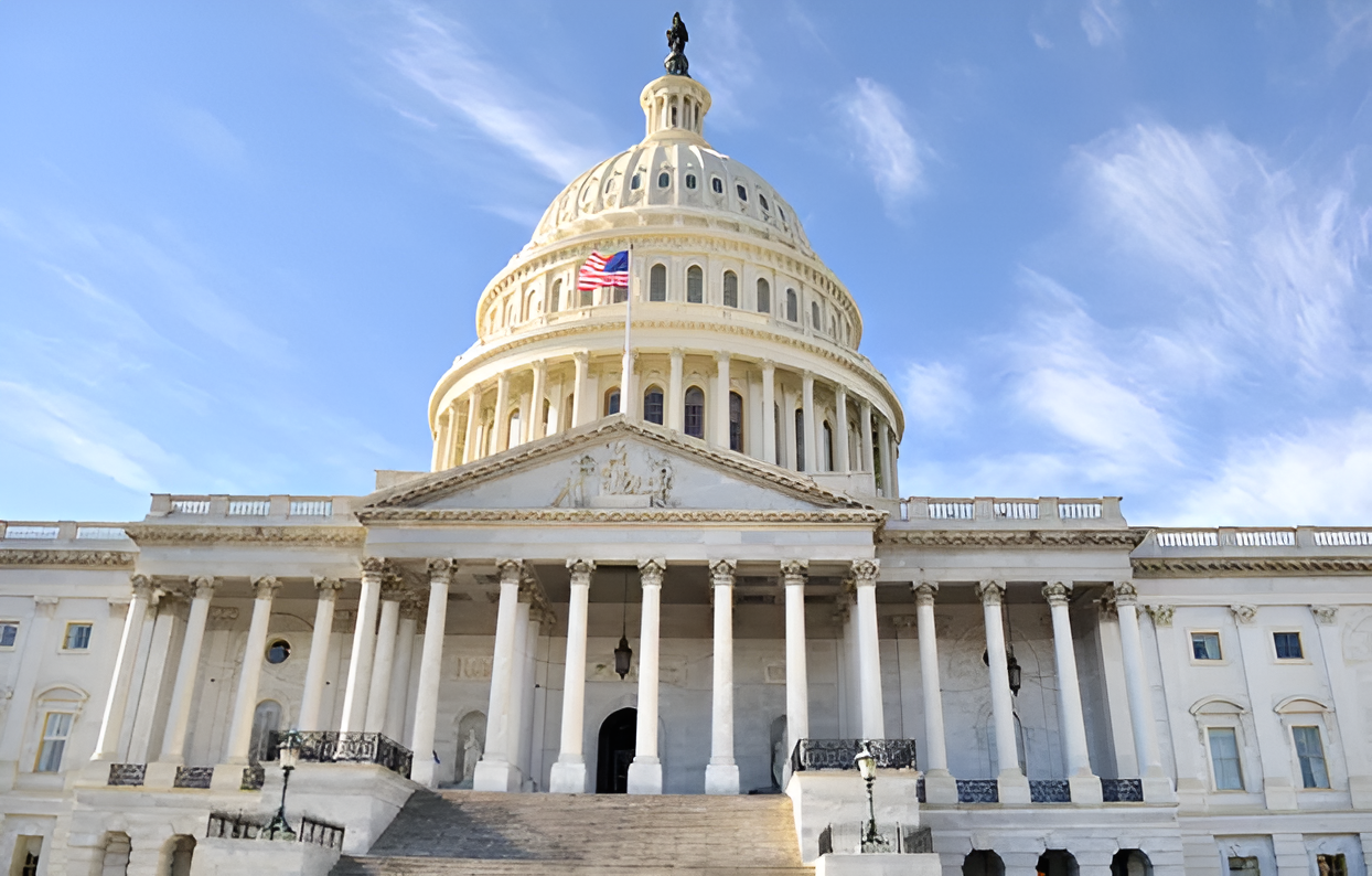 The United States Capitol building with a U.S. flag flying, viewed from the front against a blue sky with clouds.
