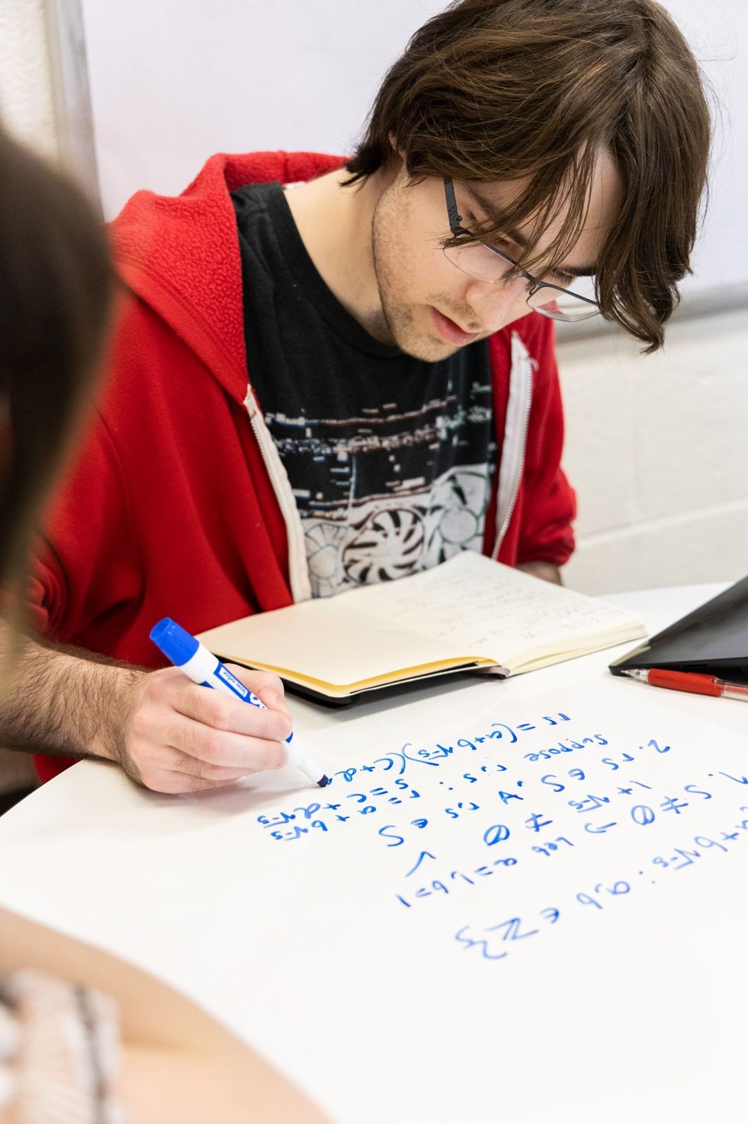 A person wearing glasses and a red hoodie writes equations with a blue marker on a whiteboard table while referencing notes.