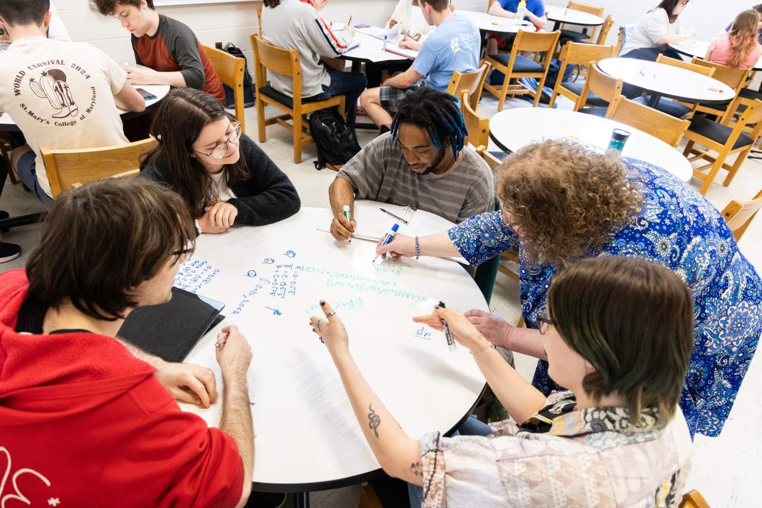 A group of students sits around a table in a classroom, writing on a whiteboard tabletop while an instructor helps them. Other groups are in the background.