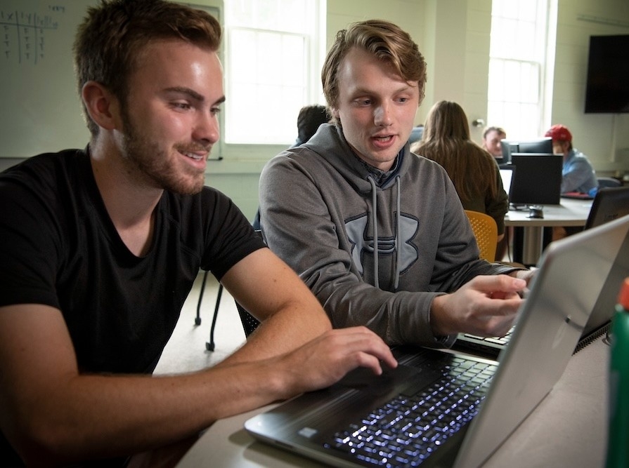 2110-1100 Two young men sit at a table working together on laptops in a classroom setting, with other students and computers visible in the background.