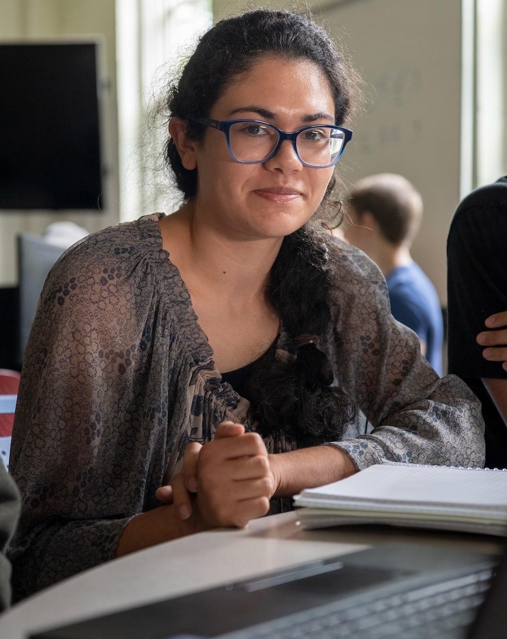 2110-1186 A person with glasses and dark hair sits at a table with a notebook, looking at the camera. Other people and a whiteboard are visible in the background.
