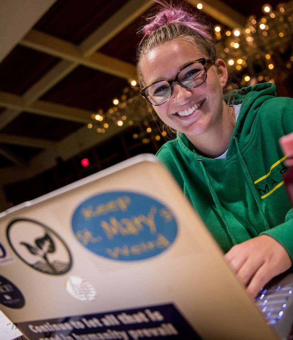 25024982837_bd4b73ff80_o A person wearing glasses and a green hoodie smiles while sitting at a table, working on a laptop decorated with various stickers.