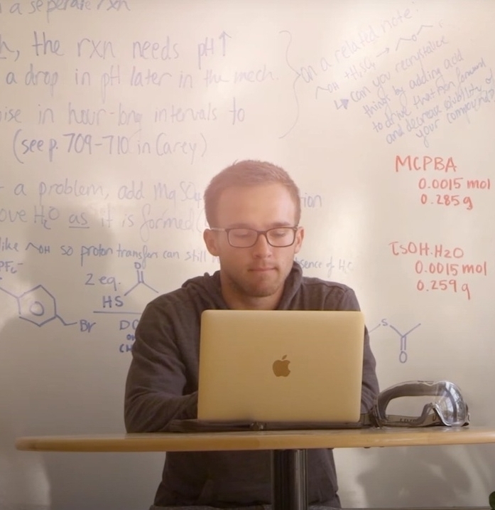 Screenshot2025-06-23at1.49.10P A man works on a laptop at a small table in front of a whiteboard covered with handwritten notes, diagrams, and chemical formulas.