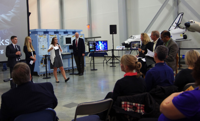 smcm-career-dev-pax-presentation A group of people present and listen in a hangar-like room with aerospace equipment and a drone on display in the background.