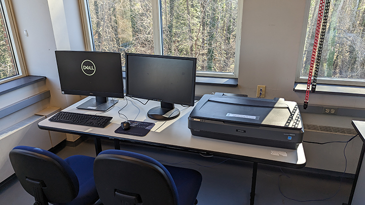 A desk with two monitors, keyboard, mouse, and a large flatbed scanner sits by windows with a view of trees. Two chairs are positioned at the desk.