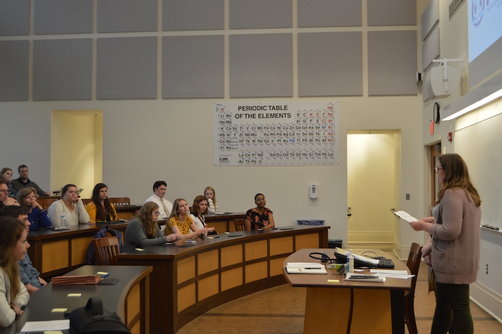 A woman stands at a lectern addressing a classroom of students seated at curved desks, with a periodic table on the wall behind them.
