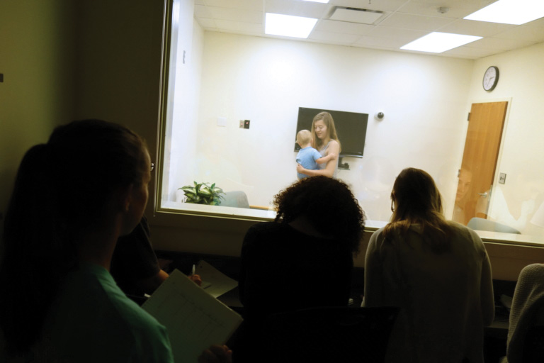 Four people observe through a window as a woman holds a baby in a brightly lit room with a TV, clock, and closed door. Some observers appear to be taking notes.