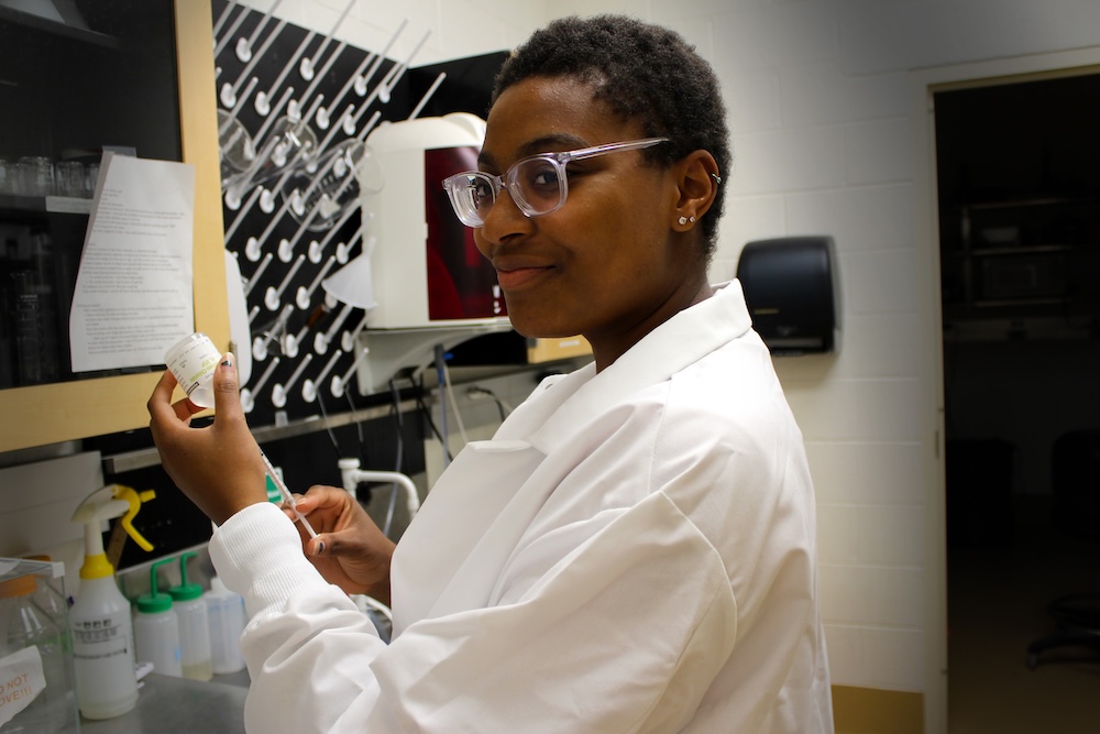 A person in a white lab coat prepares a syringe with liquid from a small container in a laboratory setting.
