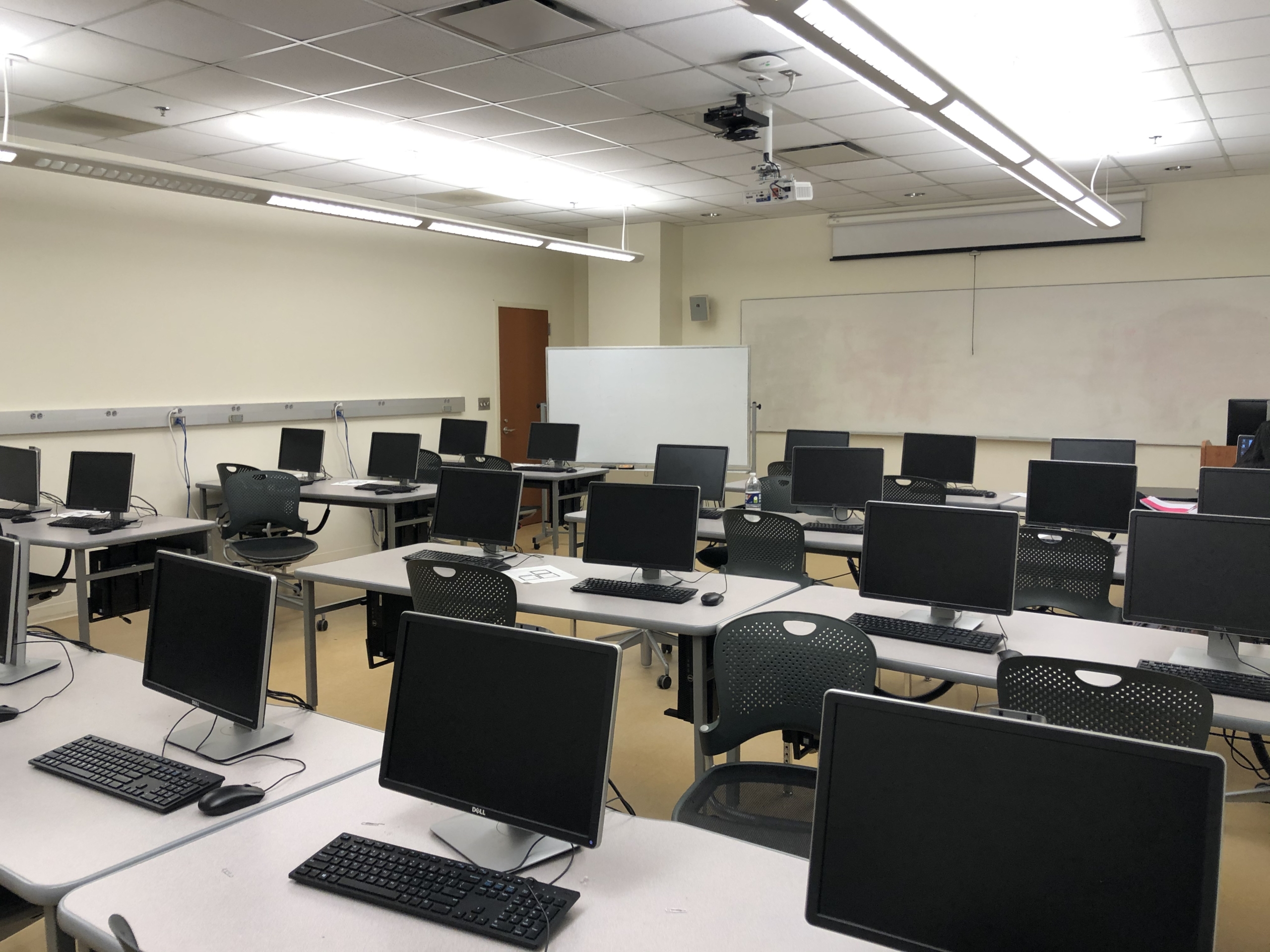 A computer lab with multiple rows of empty desks, each equipped with a monitor, keyboard, and chair, under fluorescent lighting.