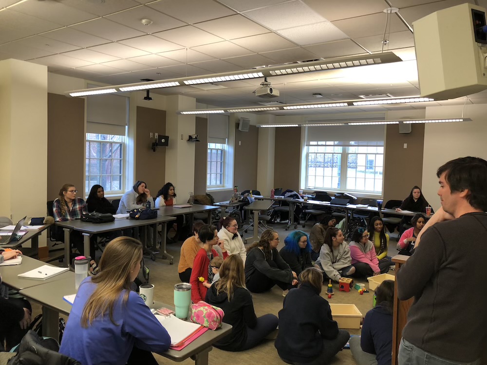 A group of students sits around desks and on the floor in a classroom, listening to a person speaking at the front near a table with colorful objects.