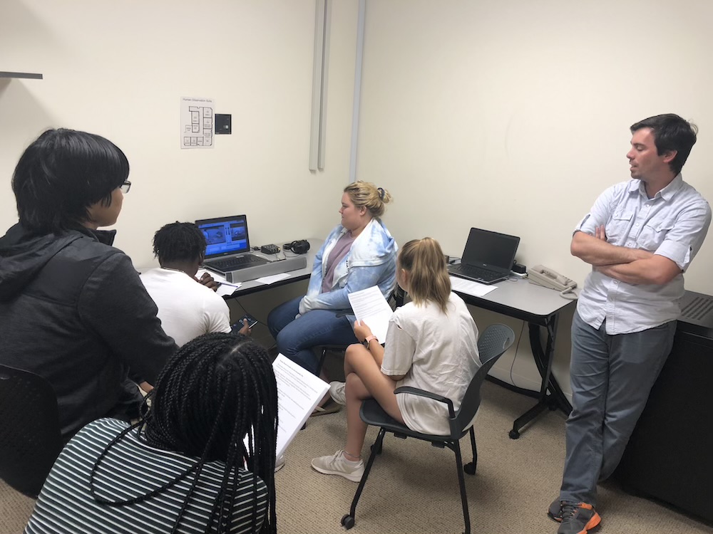 A group of students sit and take notes while a woman shows something on a computer screen; a man stands nearby with arms crossed, observing.