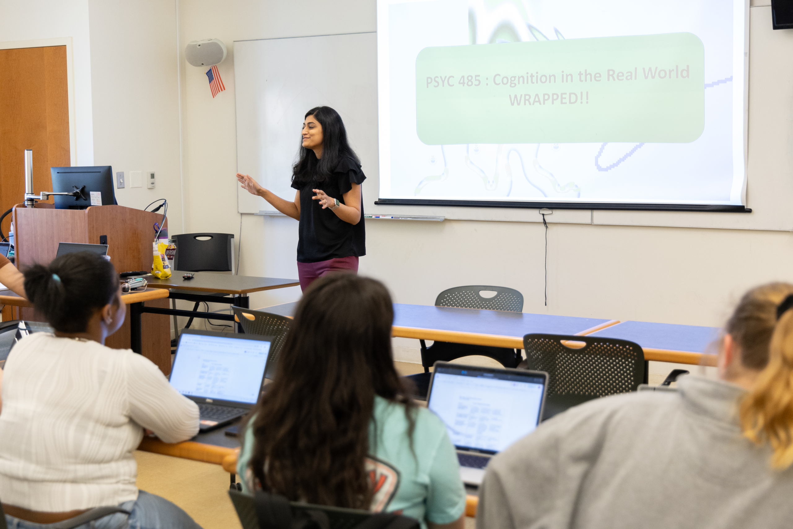 53818603327_7e3b1f1b86_o A woman stands and speaks at the front of a classroom, presenting a slide titled "PSYC 485: Cognition in the Real World WRAPPED!" to students seated with laptops.