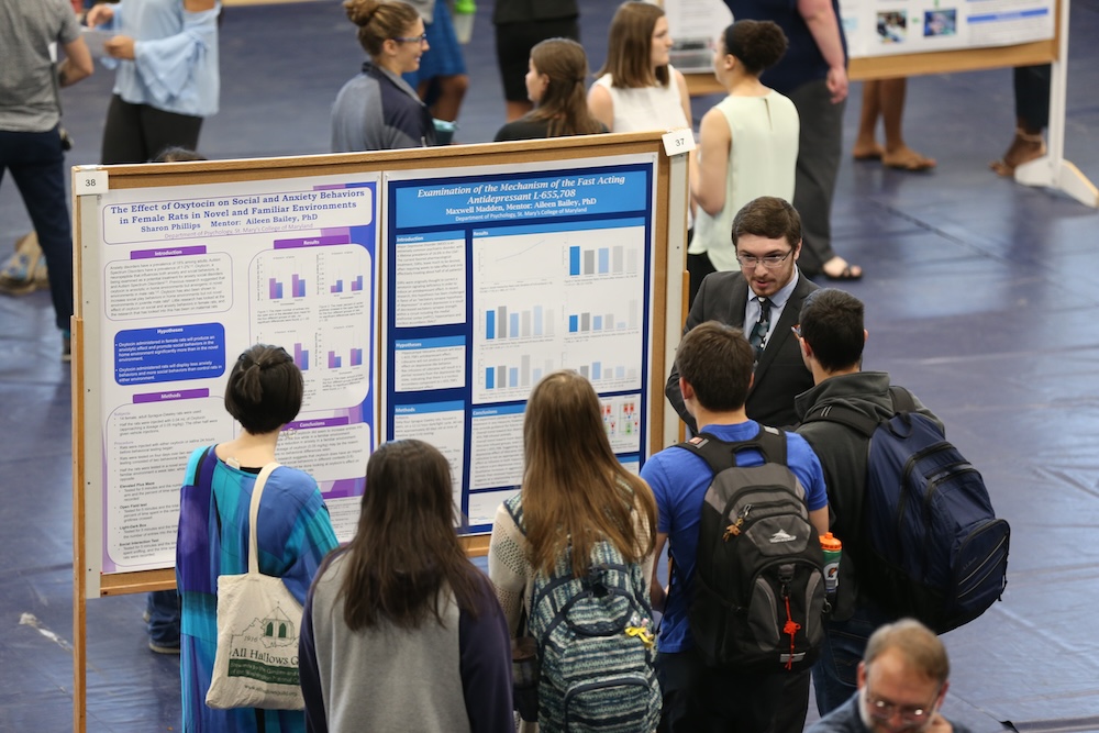 A group of people stands in front of research posters at an indoor academic conference or science fair, listening to a presenter.