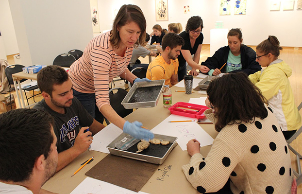 A group of students sit around a table drawing objects while an instructor wearing gloves distributes trays with specimens in an art classroom setting.