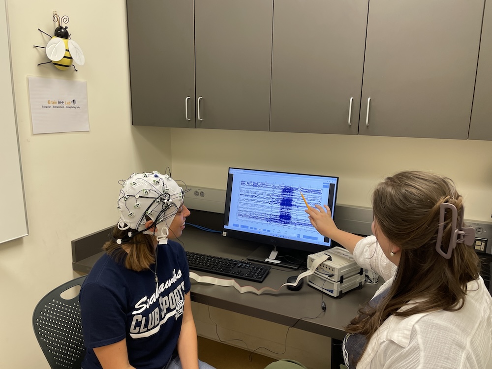 Two women sit at a desk; one wears an EEG cap while the other points at brainwave data displayed on a computer monitor in a clinical setting.