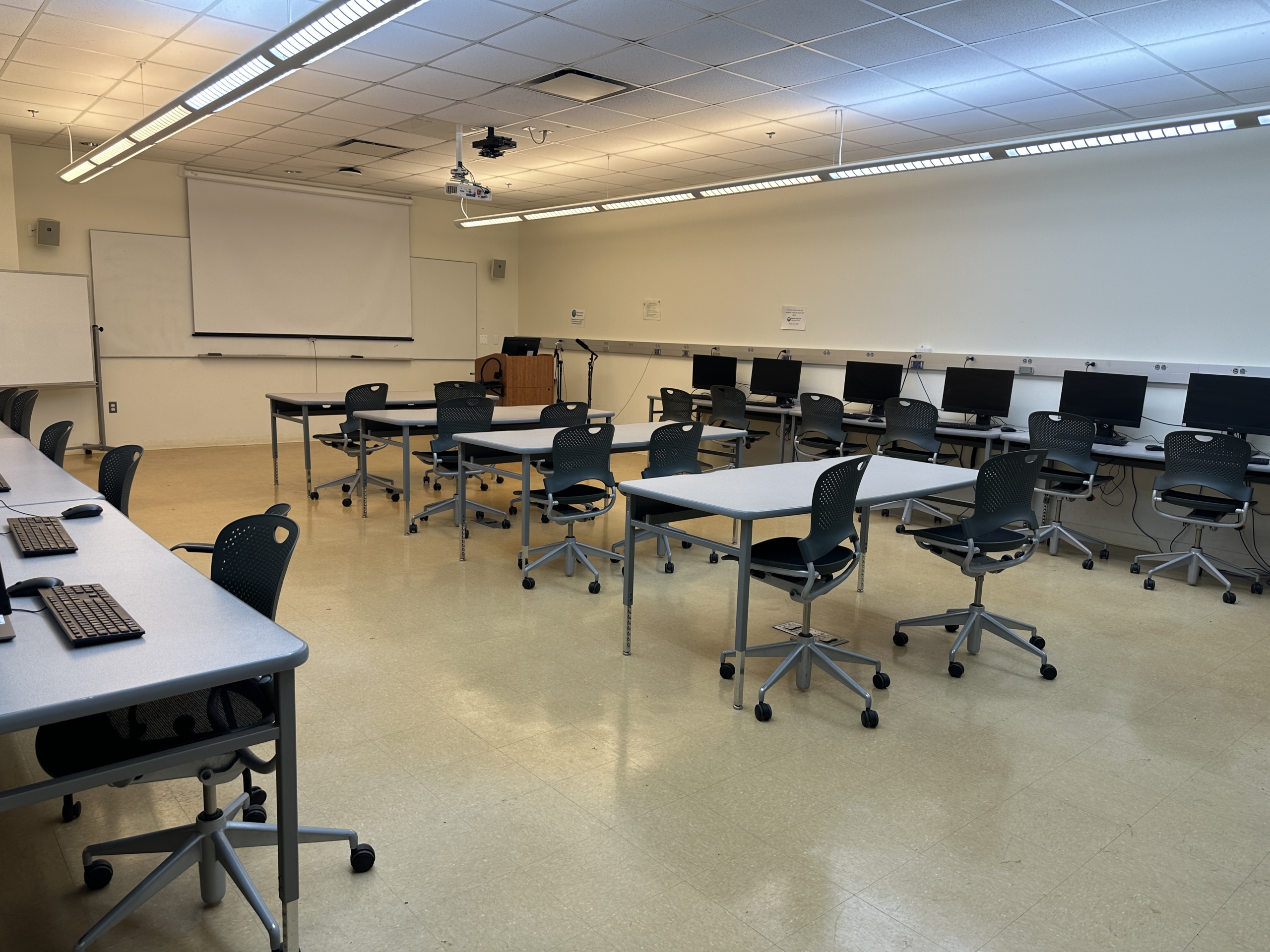 GH81NewLayout A computer lab classroom with several desks, rolling chairs, desktop computers along the walls, and a projector screen at the front.