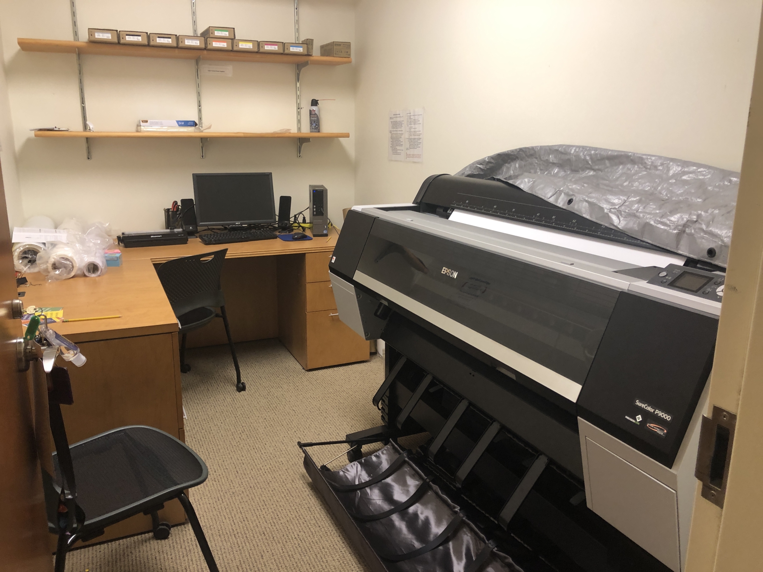 A small office contains a large Epson printer, a desk with a computer and monitor, an office chair, shelves with supplies, and rolls of paper.
