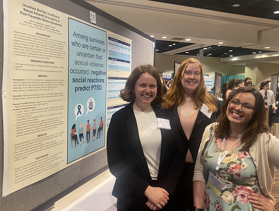 Three women stand in front of a research poster about sexual violence, social reactions, and PTSD at an academic conference.