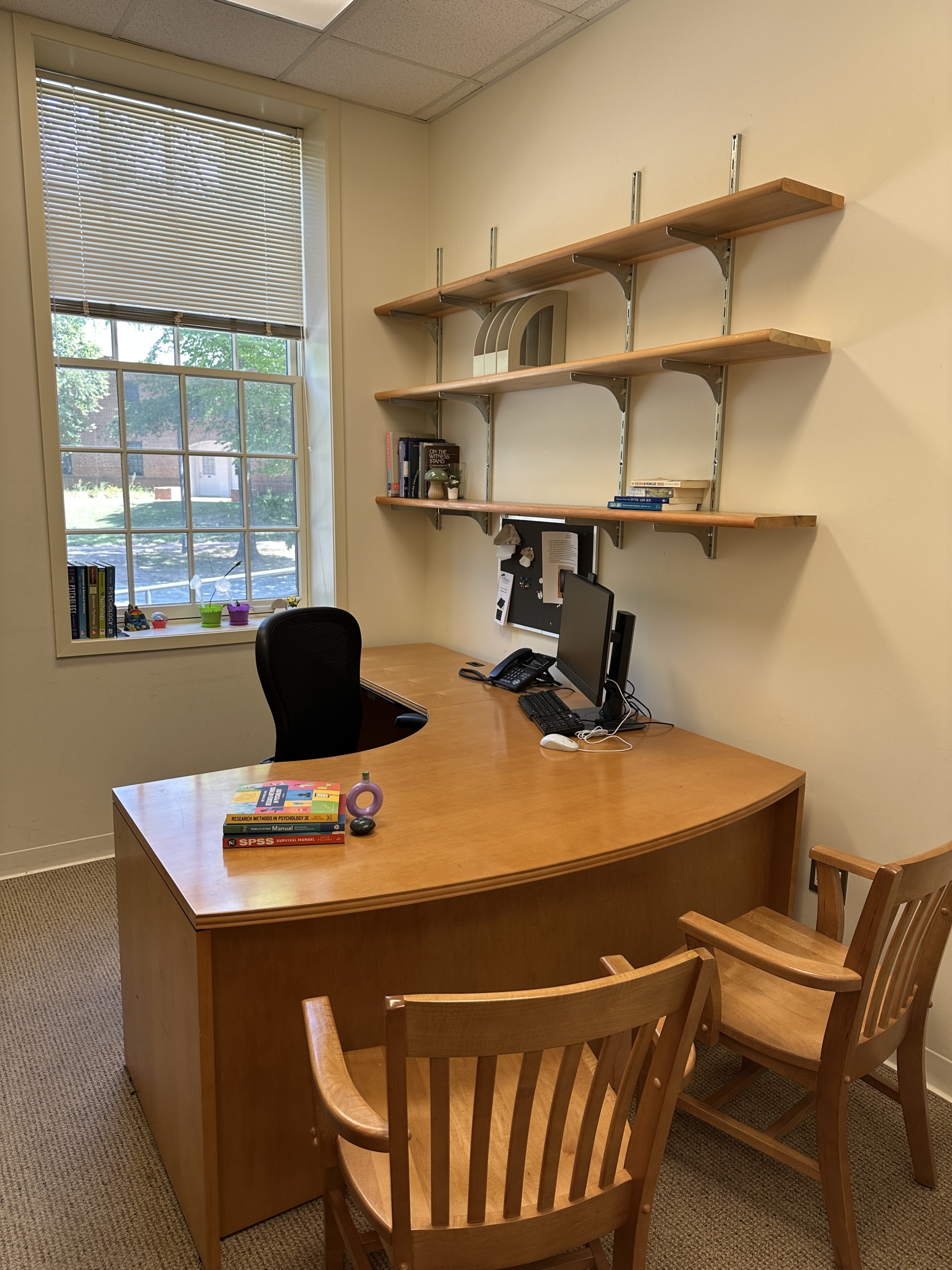 A small office with a wooden desk, black office chair, two wooden guest chairs, a computer, books, and shelves on a cream-colored wall near a window.