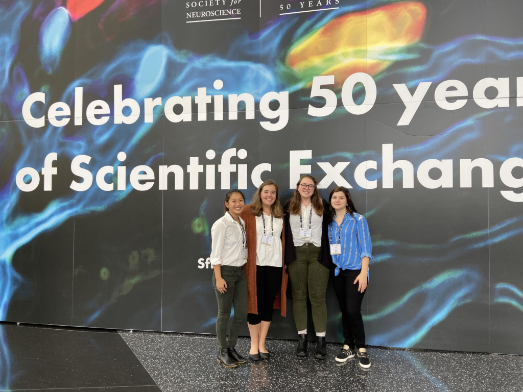Four women stand together in front of a large sign that reads “Celebrating 50 years of Scientific Exchange” at a neuroscience conference.