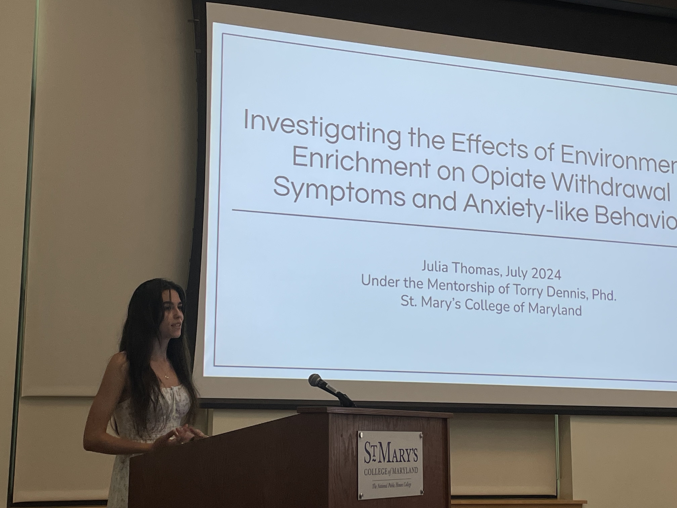 A woman stands at a podium presenting a research project titled "Investigating the Effects of Environmental Enrichment on Opiate Withdrawal Symptoms and Anxiety-like Behavior" at St. Mary's College of Maryland.