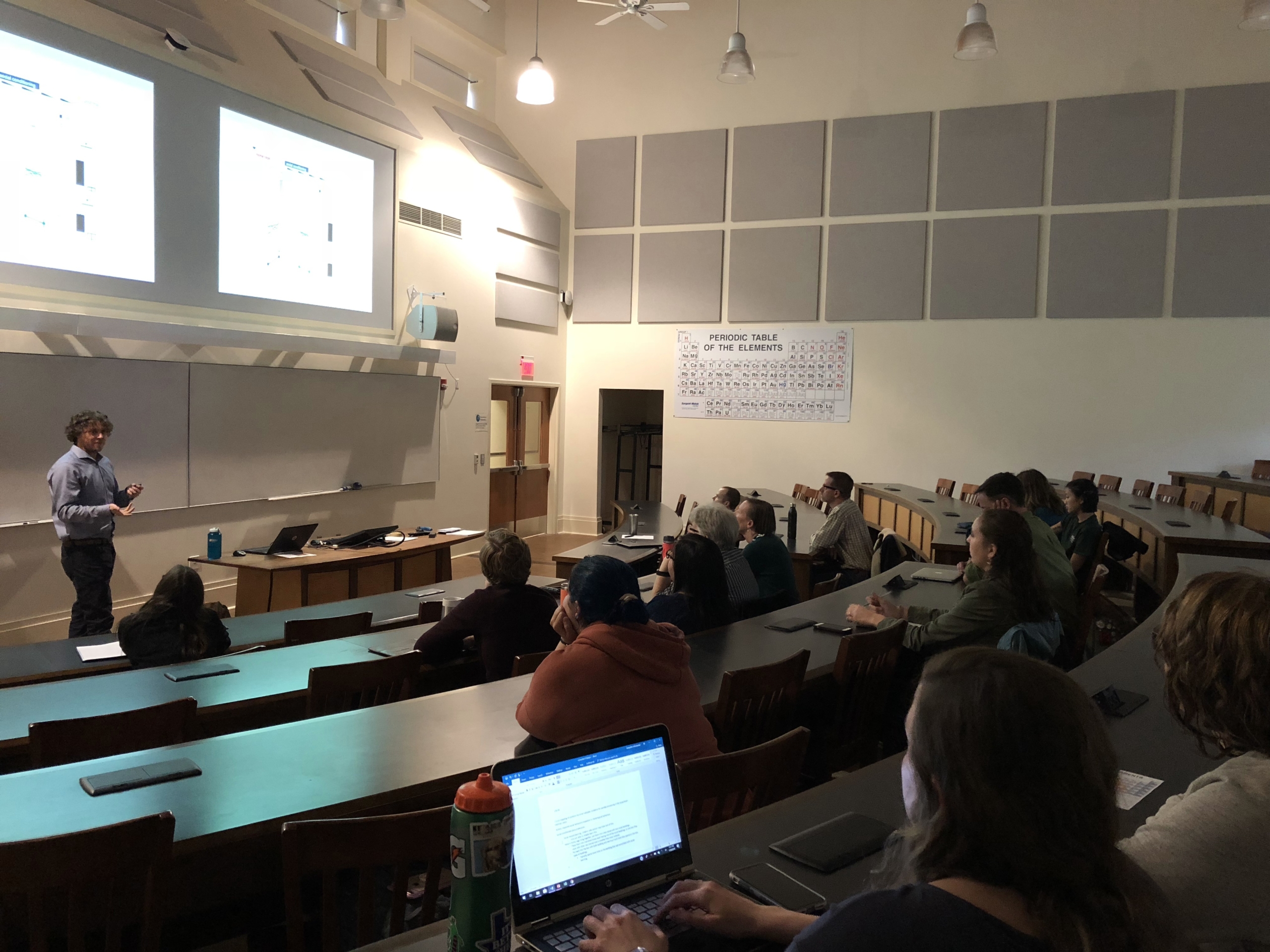 EastmanLewis08_1118.jpg A professor presents slides to students in a college lecture hall. Some students take notes on laptops, while others listen. A periodic table is displayed on the wall.