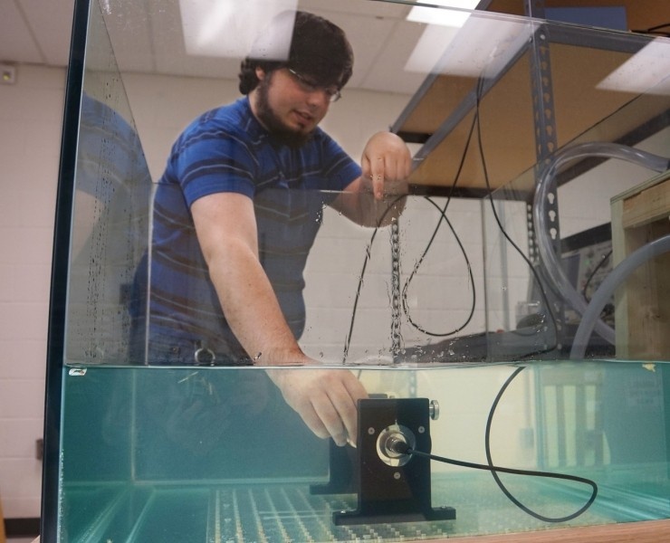 slider1-740x600_c 2 A person in a blue striped shirt adjusts equipment inside a large water tank in a laboratory setting.