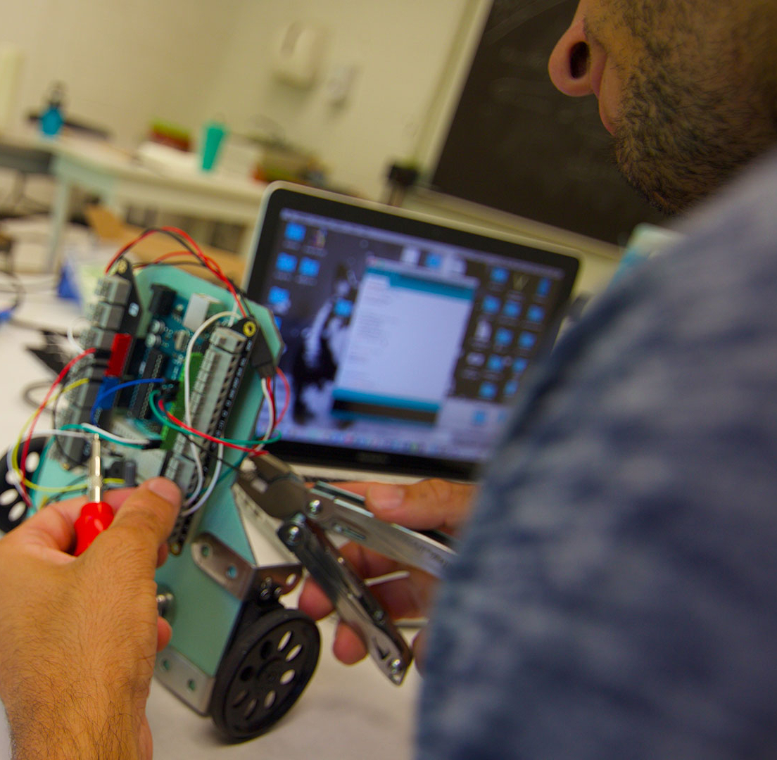 smcm-science-smp-robot-cirillo A person works on wiring a small robot in front of a laptop displaying technical software on a cluttered desk.