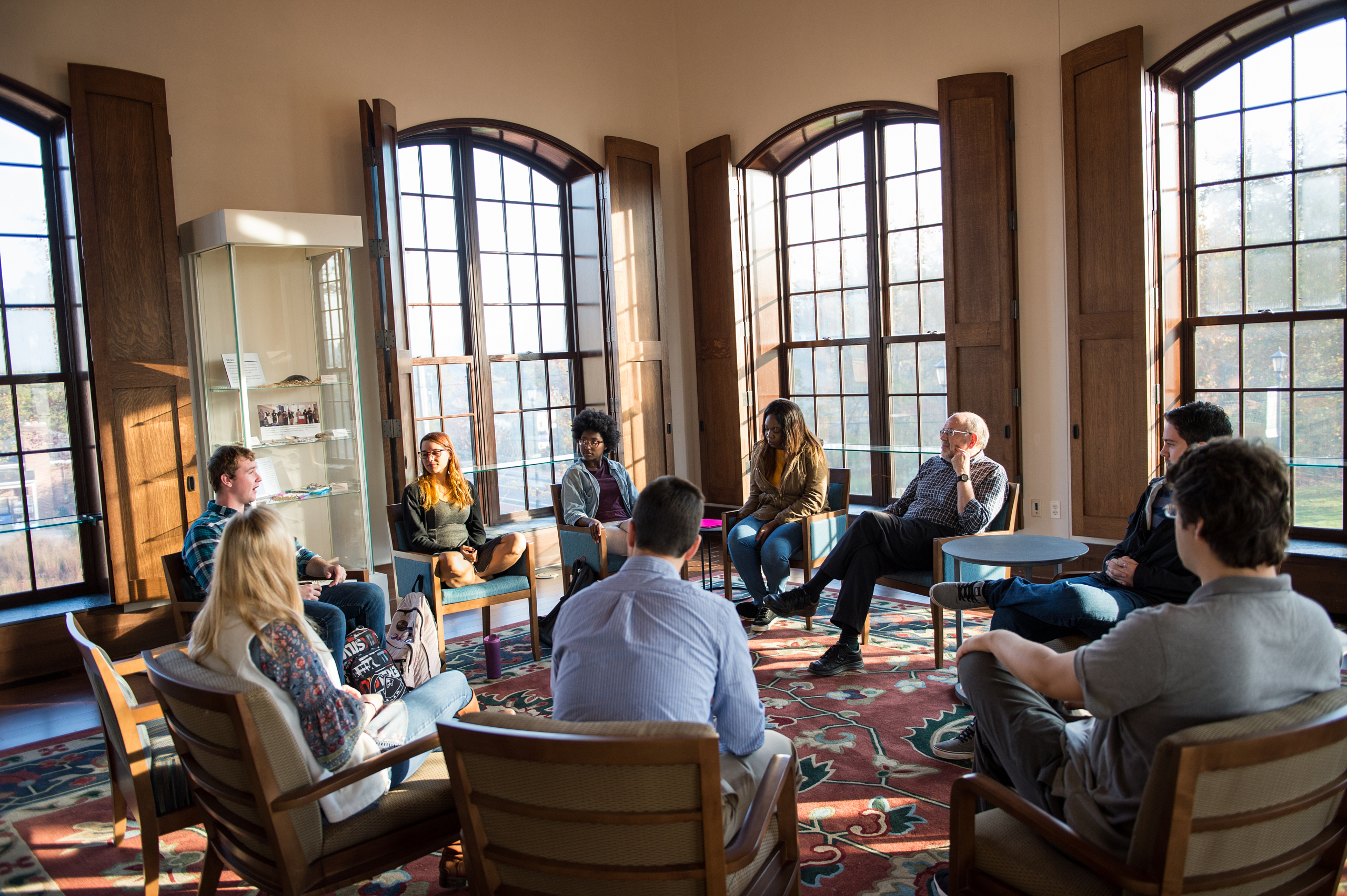 SMCM_11-3-16_0343 A group of people sit in a circle in a sunlit room with large windows, engaged in a discussion.