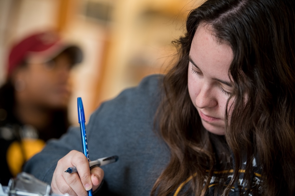 A person with long brown hair writes with a blue pen while looking down, with another person blurred in the background.