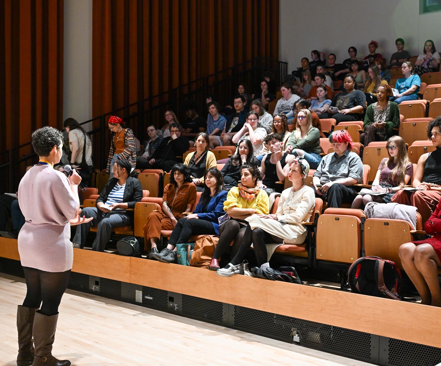 54407318971_1e50e14c03_k A person stands and speaks into a microphone in front of a lecture hall audience seated in tiered rows, listening attentively.