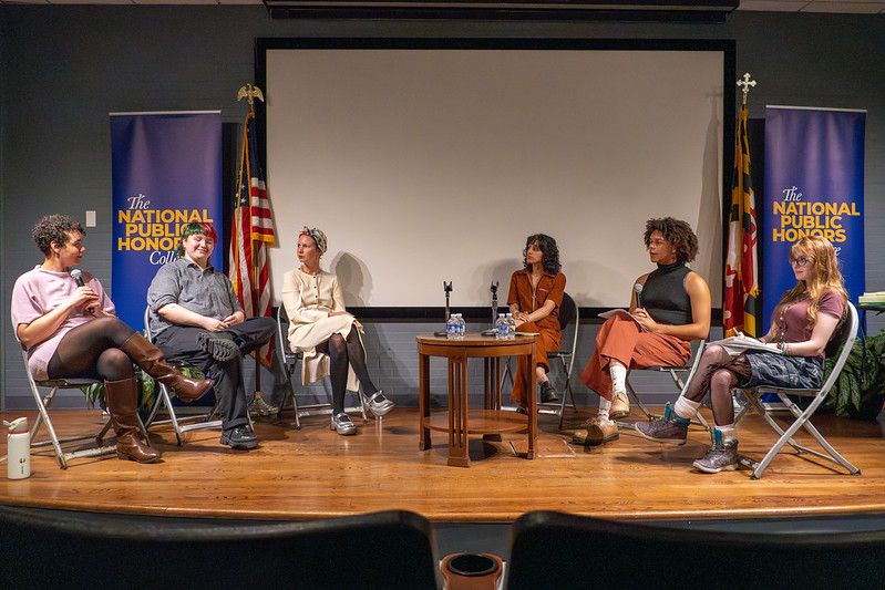 Six people sit in a panel discussion on stage in front of a screen, flanked by two "National Public Honors College" banners and flags.
