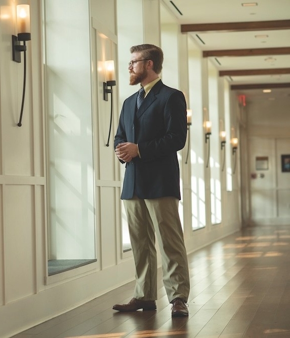 2110-1991 A man in a suit stands in a hallway with wooden floors, looking out of large windows with both hands clasped in front of him.
