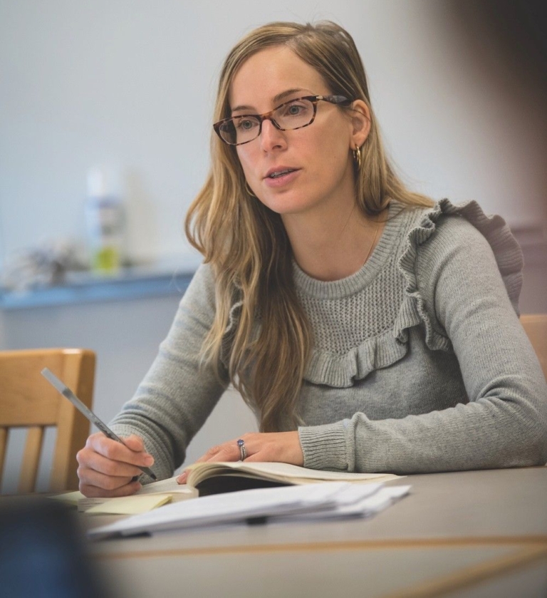 38886392925_a7f6994c1d_o A woman with glasses and long hair sits at a table, holding a pen and looking attentive, with a notebook open in front of her.