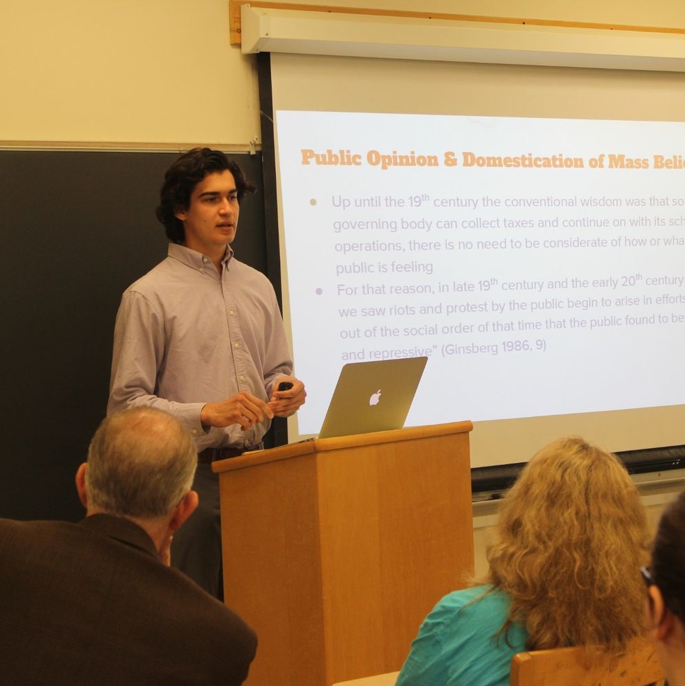st_marys_project_2017 A person stands at a podium giving a presentation in a classroom, with a slideshow on public opinion and mass belief displayed behind them.