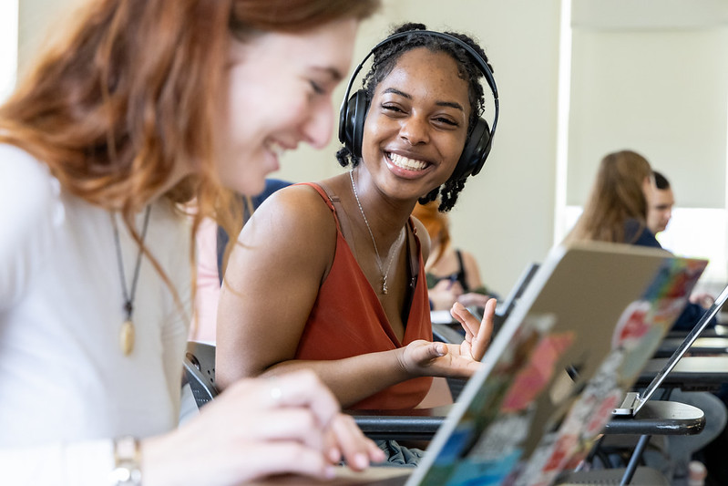 Two students sit at desks using laptops; one smiles and wears headphones, while the other focuses on typing. Other students are visible in the background.
