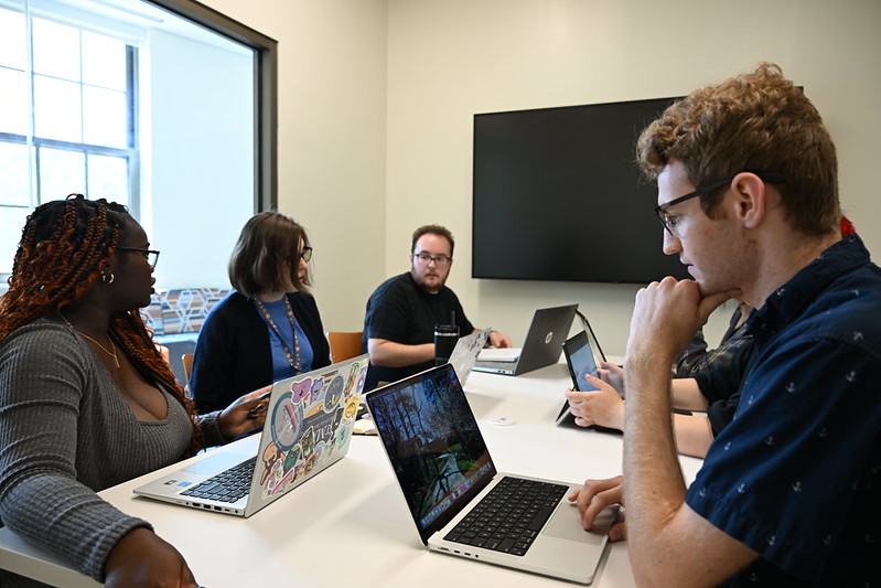 Five people sit around a table with laptops in a meeting room, engaged in discussion. A large blank screen is mounted on the wall behind them.