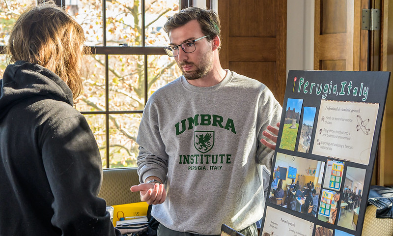 Photo by Chuck Steenburgh A man in an "UMBRIA INSTITUTE" sweatshirt discusses a display board about Perugia, Italy with another person at an indoor event.