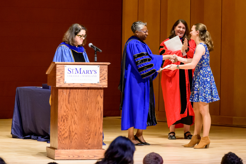 picture of student on stage being presented with an award by President Jordan with person in the background clapping