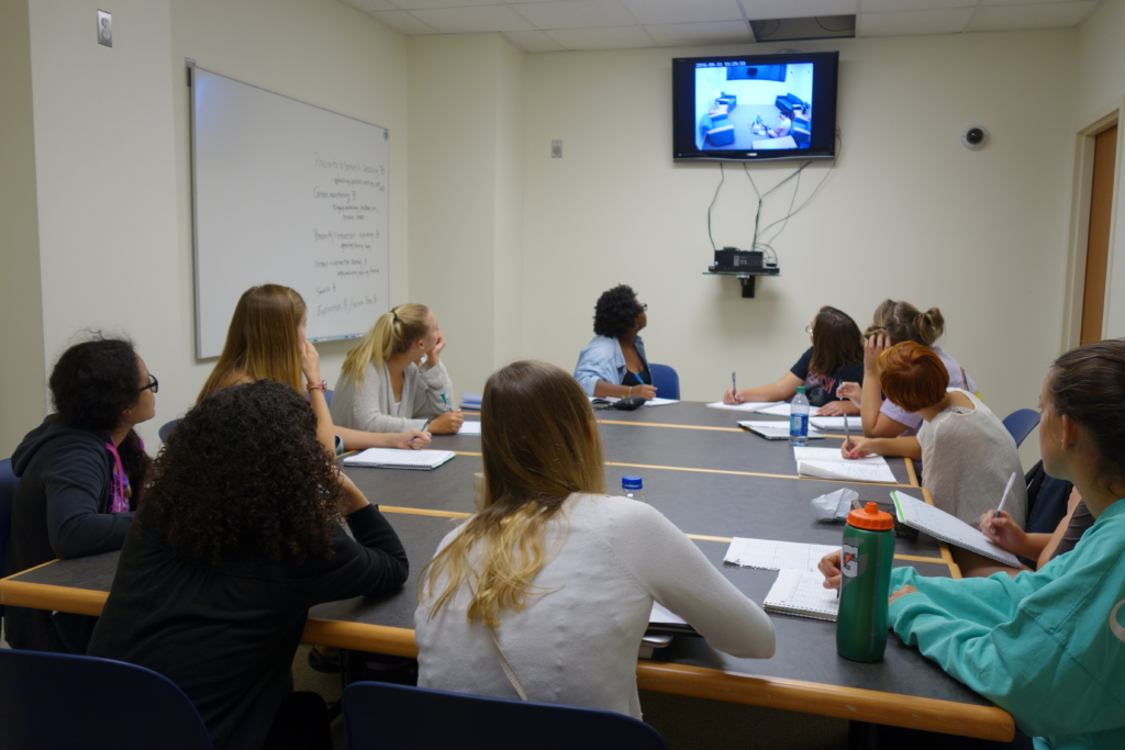 students siting asround a table watching an observation video on the TV and taking notes