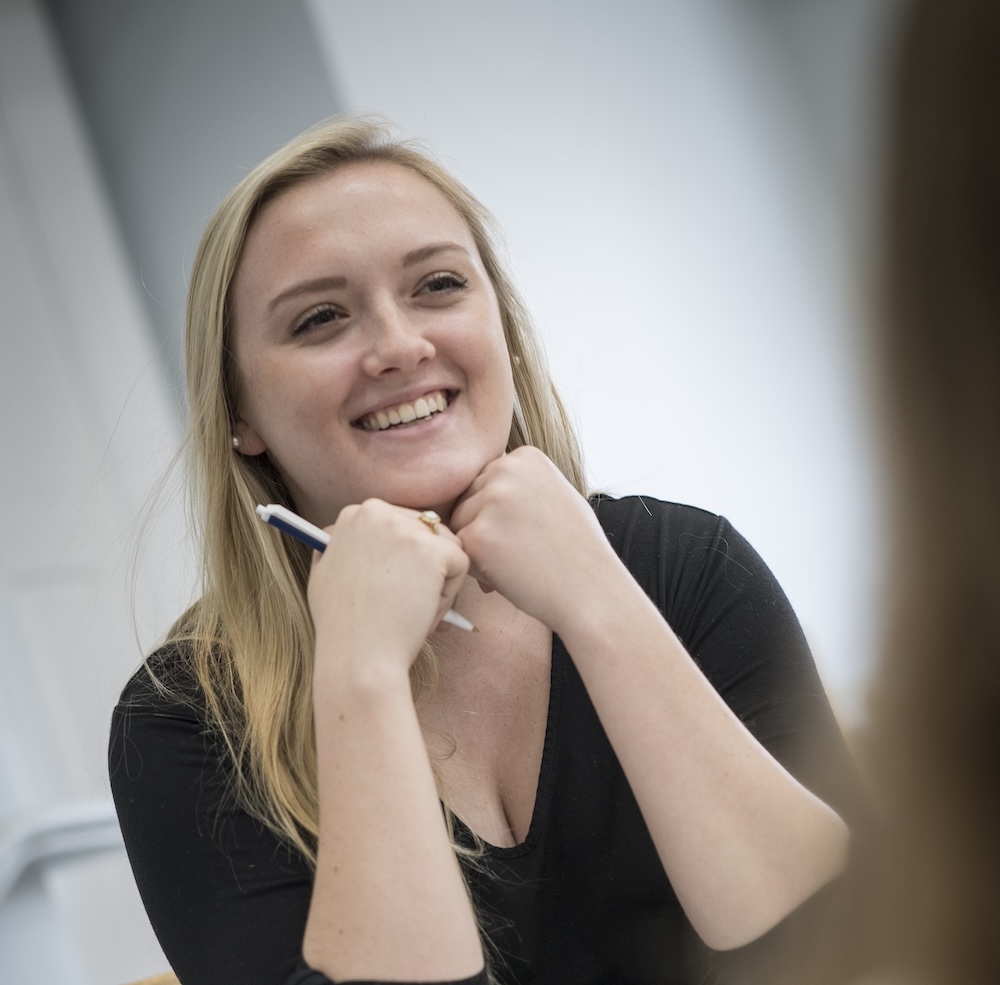 2131-1983 A woman with long blonde hair sits at a table, smiling and holding a pen, resting her chin on her hands while listening to someone off camera.